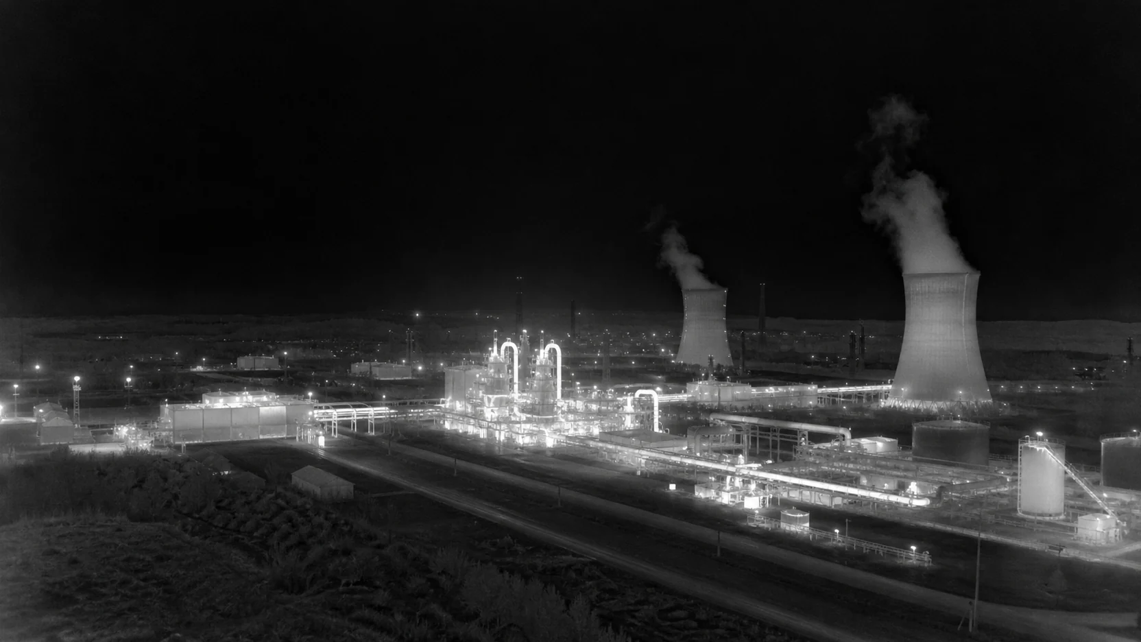 Monochrome white-hot thermal image of an industrial plant at night: cooling towers, pipework and tanks stand out bright against a cold background