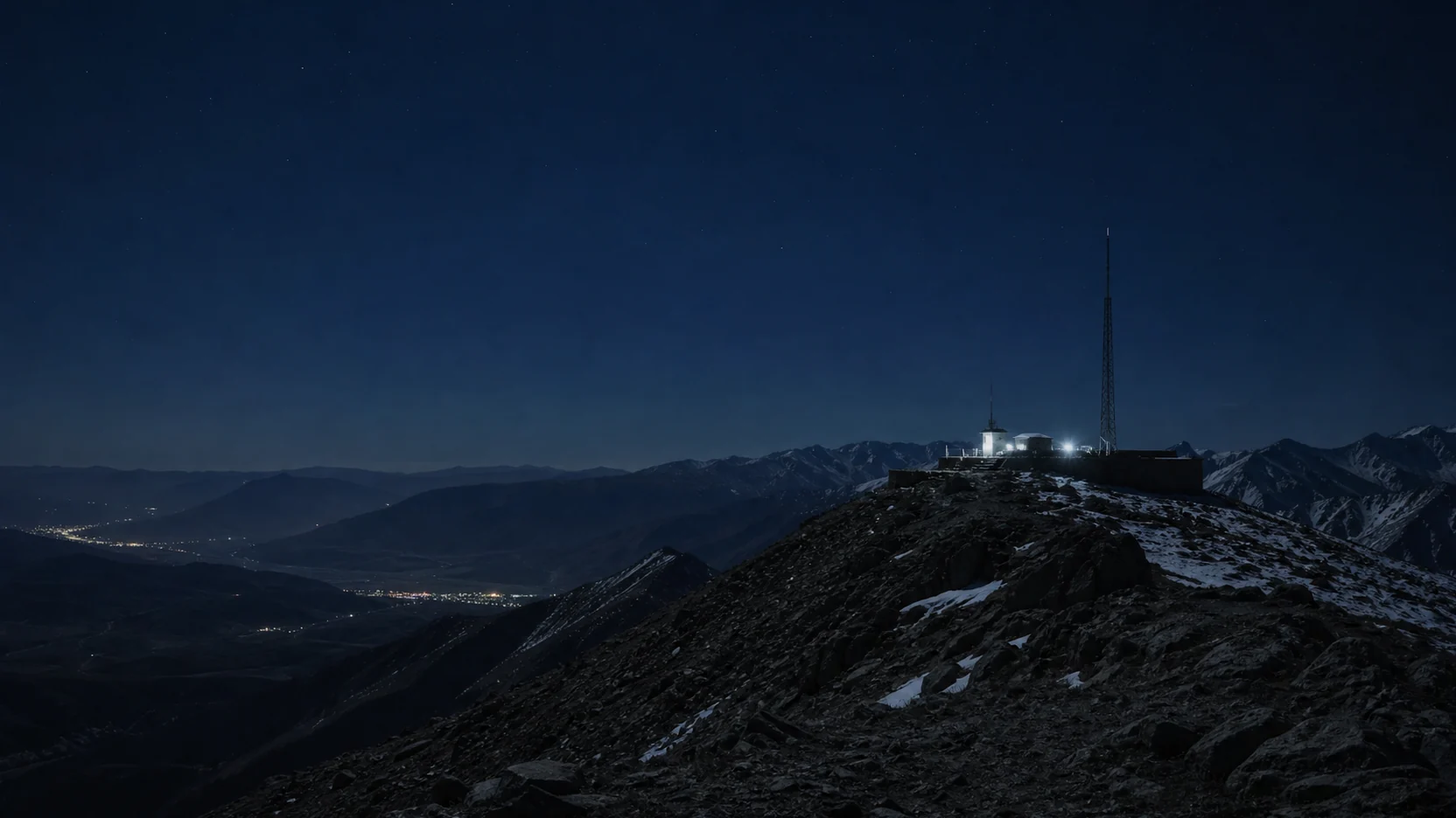 A floodlit shelter and a thin mast on a remote mountain crest above a dark valley with scattered settlement lights under a faint star field