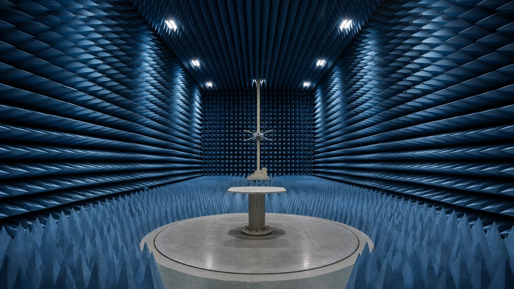 Blue pyramidal RF absorbers converging around a central turntable and a distant measurement antenna inside an anechoic chamber
